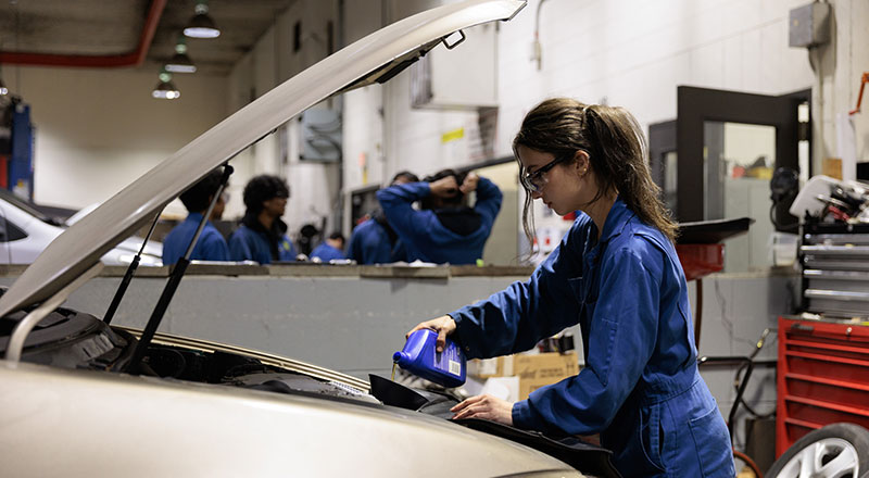 Person in blue coveralls pours motor oil into a vehicle with the hood open in an automotive workshop, while other students work in the background.