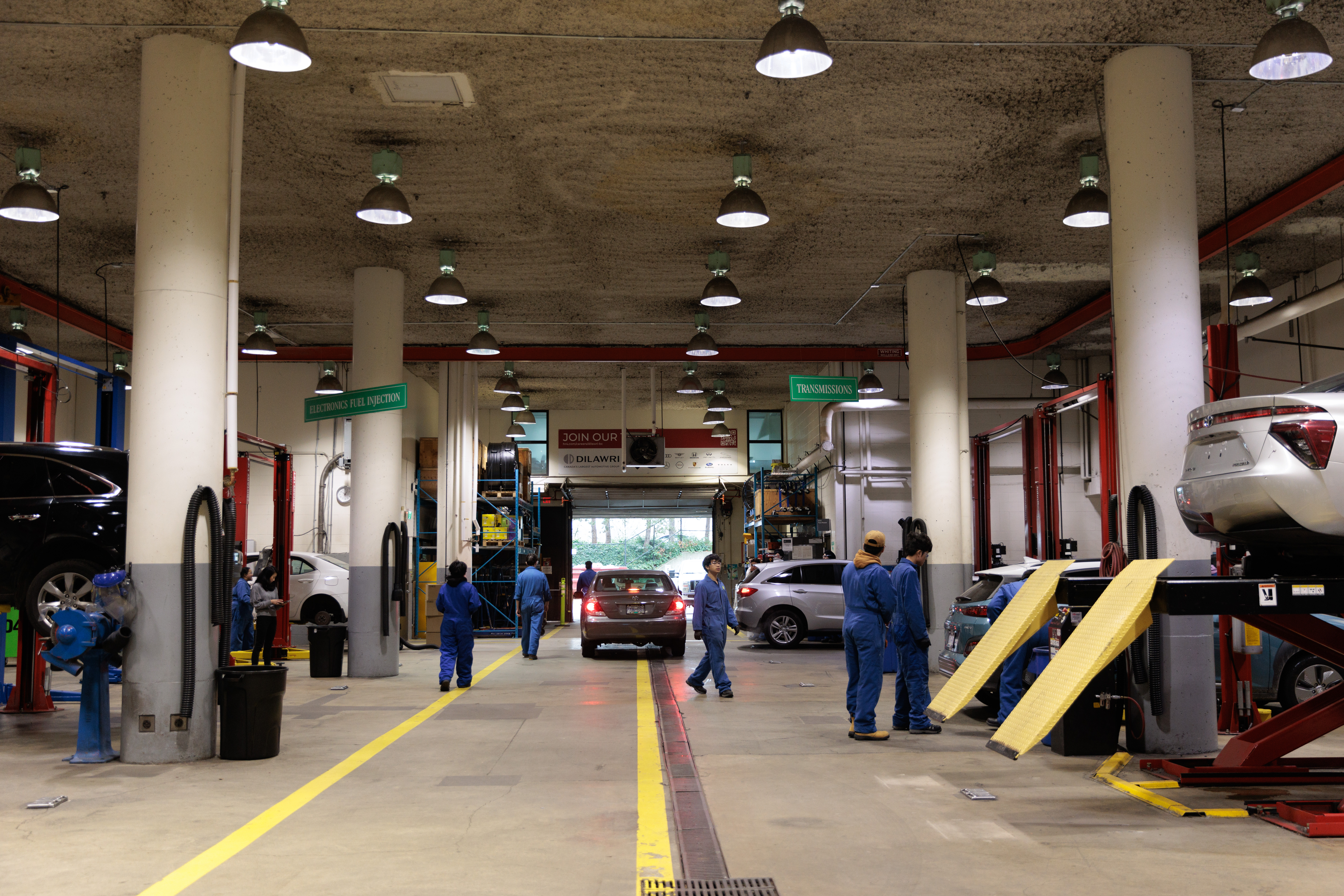 Wide view of a busy automotive service workshop with multiple vehicles being repaired. Several students or technicians in blue coveralls work around cars on lifts and in service bays. A vehicle drives down the center aisle marked with yellow floor lines, while overhead industrial lights illuminate the large garage. Signs for “Electronics Fuel Injection” and “Transmissions” hang above the work area