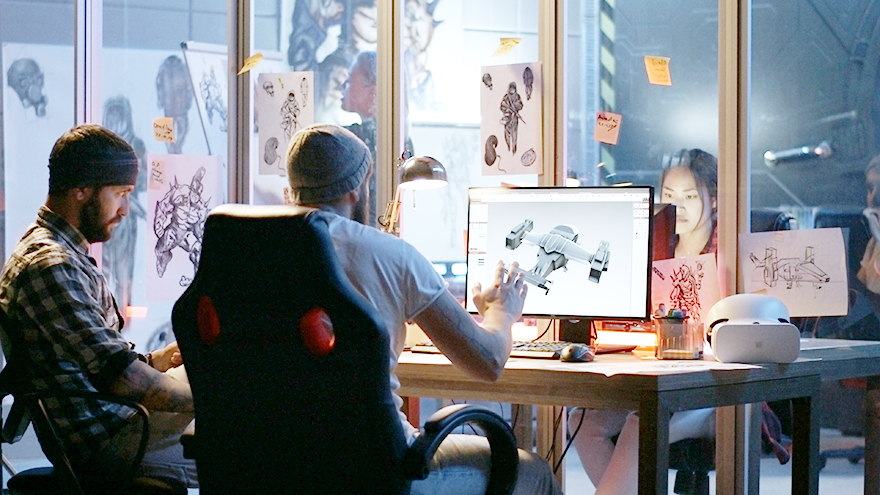 A group of people work together in a creative studio filled with concept art sketches pinned to glass walls. Two team members sit at a desk reviewing a 3D vehicle model displayed on a computer monitor. The workspace includes drawing materials, notes, and a VR headset, suggesting a design or animation project in progress.