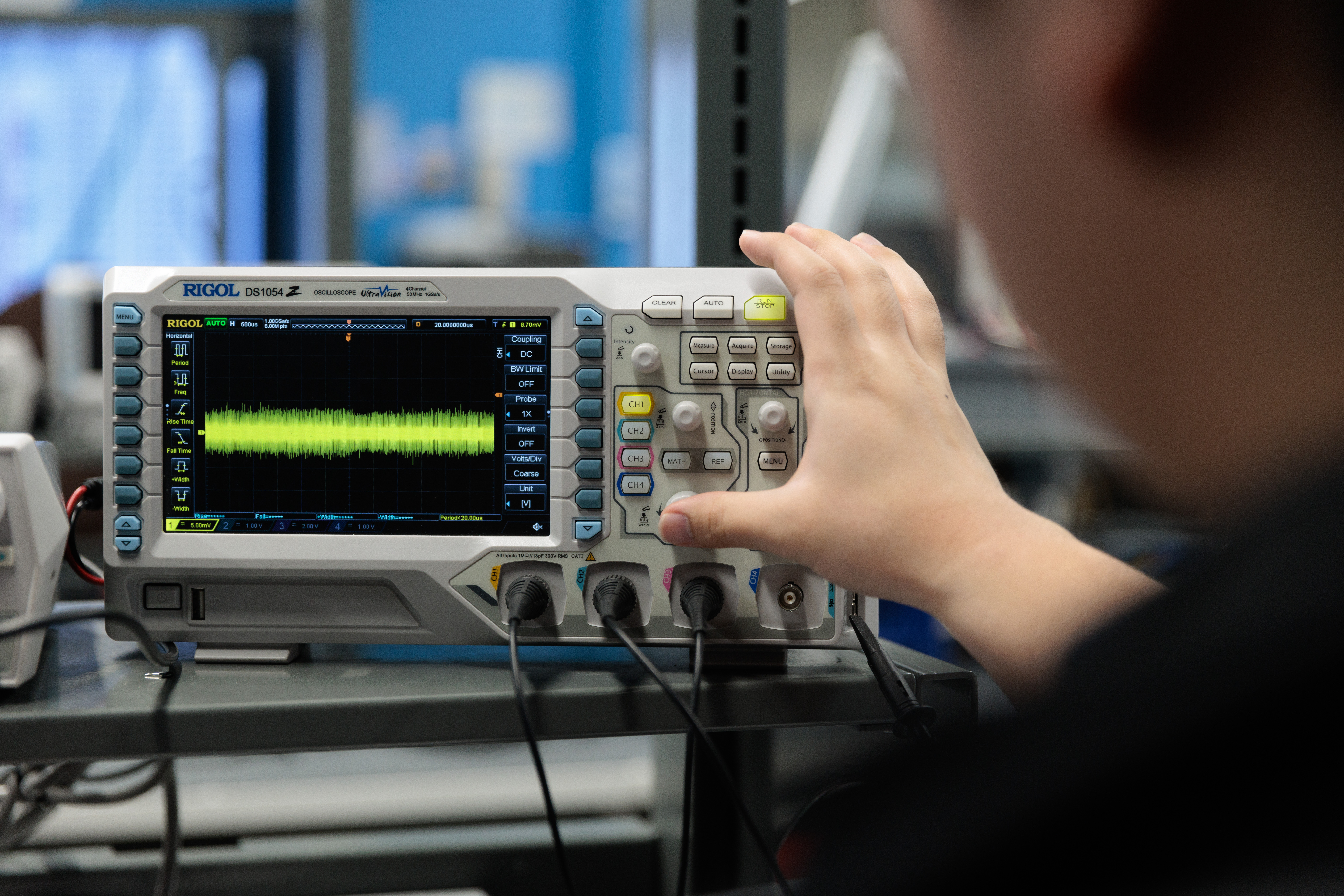 Close-up of a digital oscilloscope on a workbench displaying a green waveform signal, with several connected probes and cables. A person’s hand adjusts the control knobs on the right side of the device. The oscilloscope is labeled “Rigol DS1054Z,” and the background shows a blurred electronics lab setting.
