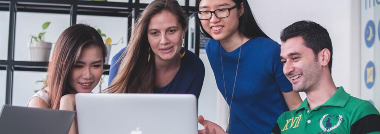 4 students of different backgrounds are looking at a laptop screen (not visible). All are smiling at what they see on the screen.