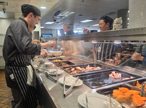 Chef serving various Asian dishes at a buffet-style food counter with trays of prepared meals.