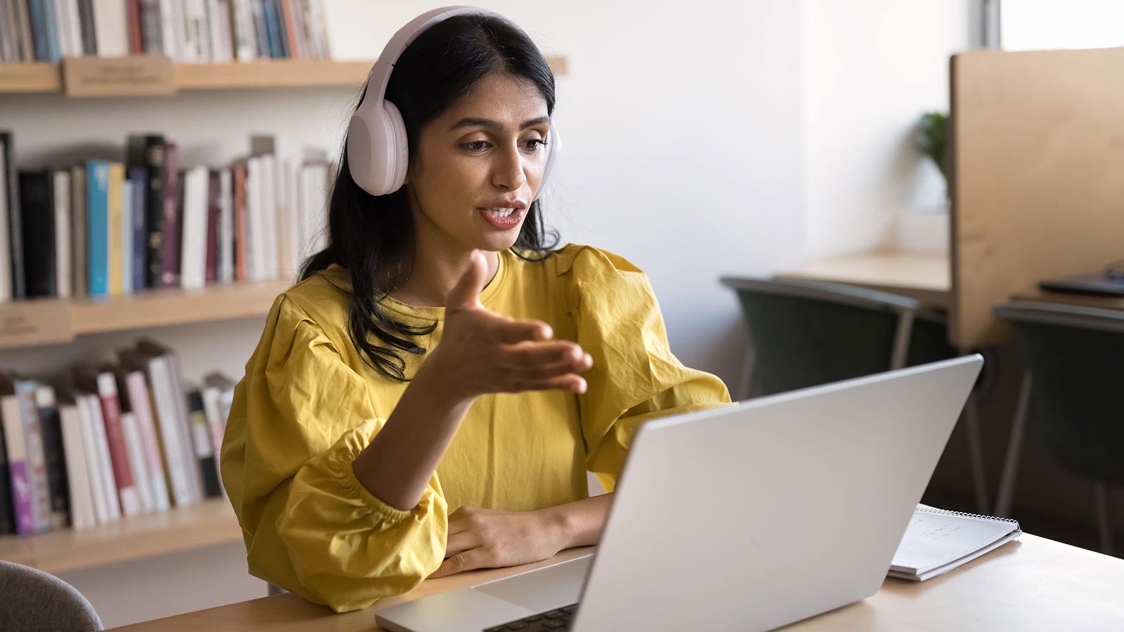 Woman wearing headphones sits at a desk in front of a laptop, speaking and gesturing during an online meeting or class, with bookshelves in the background.