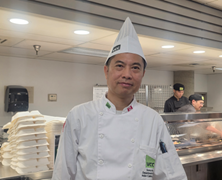 Chef in a white uniform and tall hat standing in a modern kitchen with trays stacked in the background.