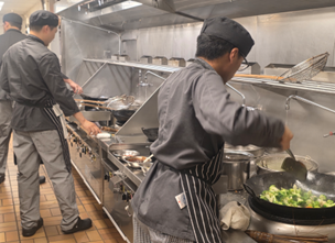 Chef serving various Asian dishes at a buffet-style food counter with trays of prepared meals.