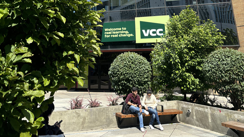 Exterior view of the VCC building, featuring a green sign with the VCC logo and the text "Welcome to real learning, for real change.". Two people are sitting on a bench in the foreground, with green trees surrounding the area.