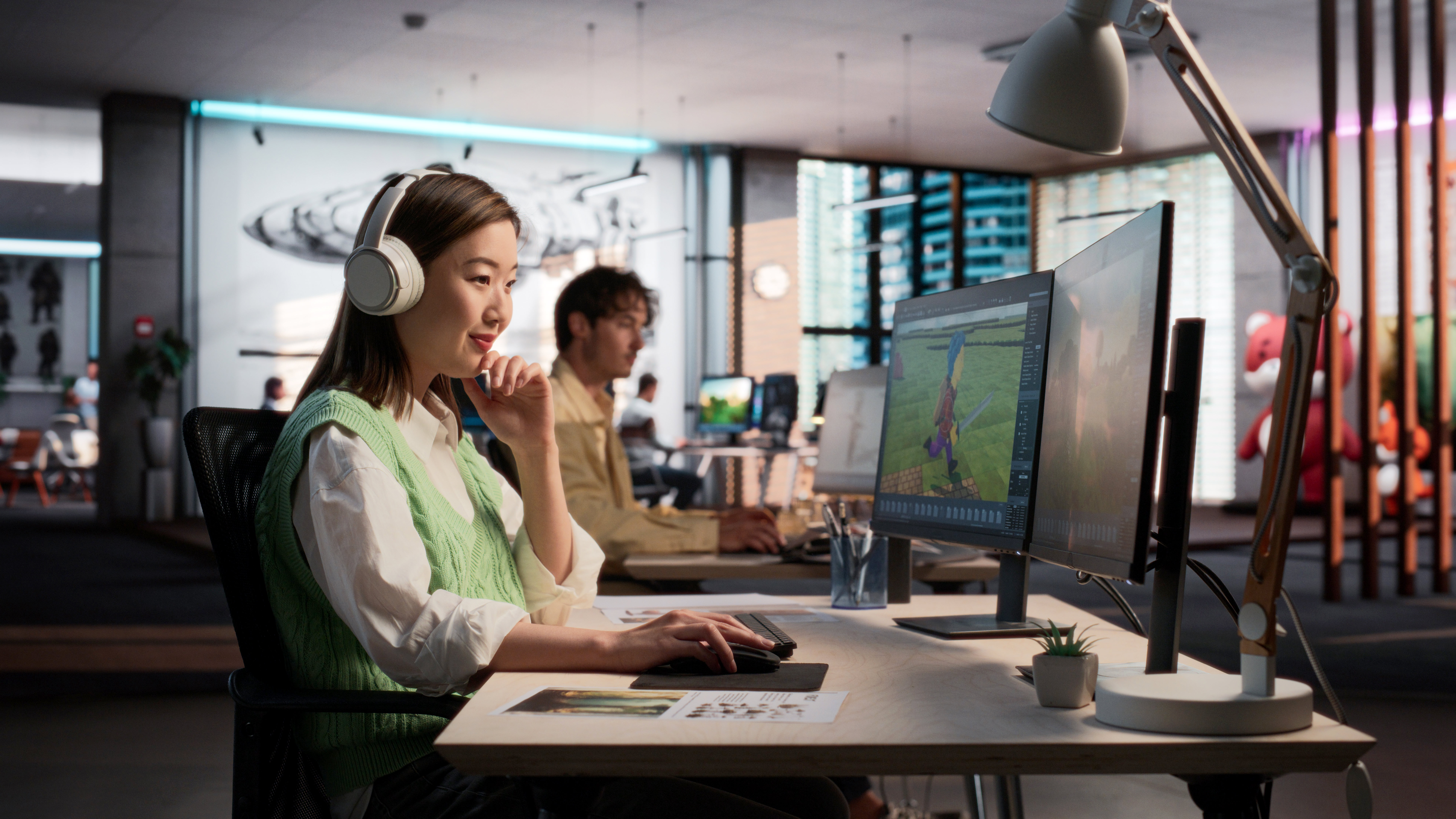 A focused woman wearing headphones edits a colorful digital project at a dual-monitor workstation in a modern, open-plan creative office.