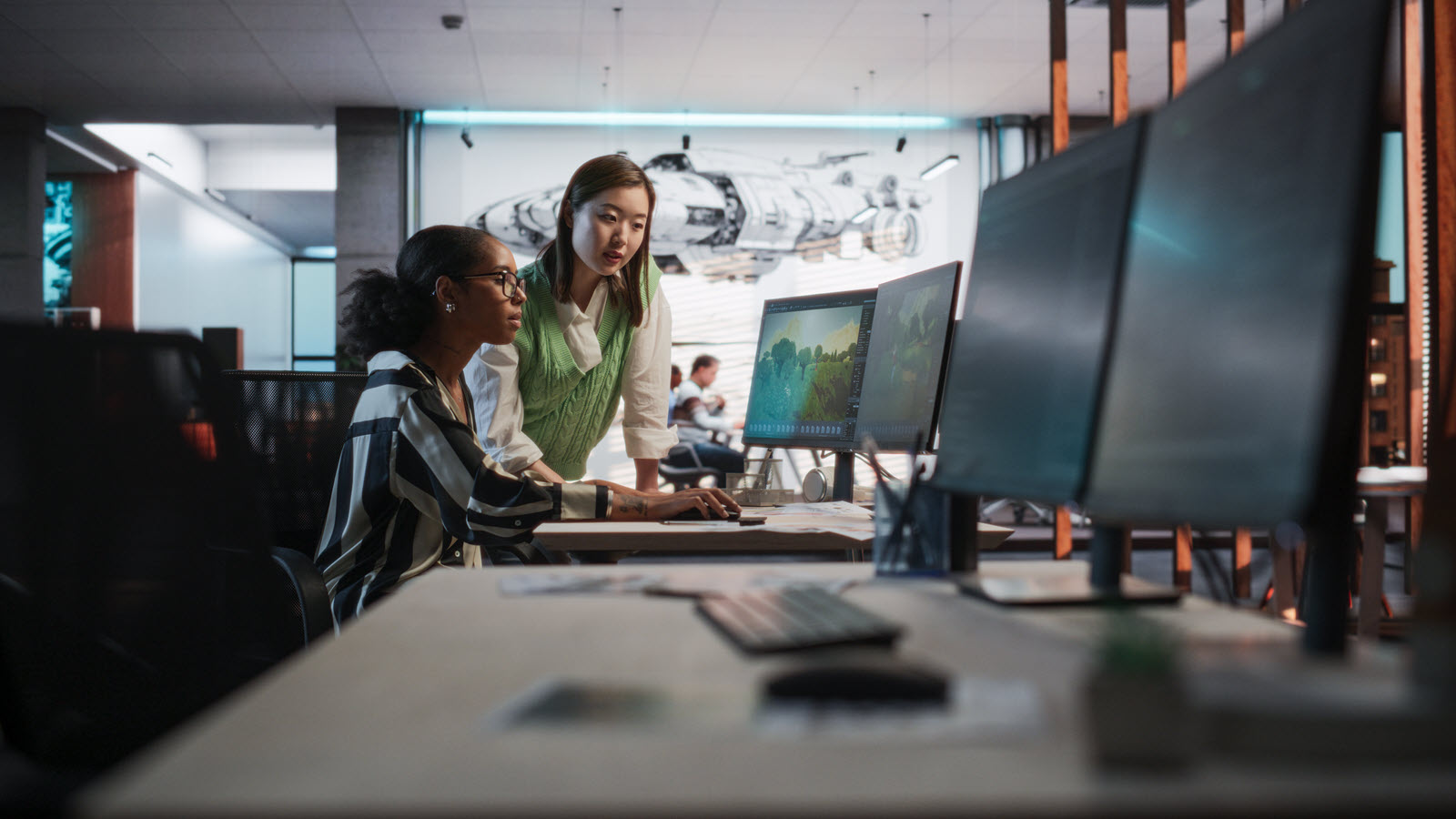 Two women collaborate at a computer workstation in a modern office, reviewing a colorful screen among large monitors; their teamwork feels focused and supportive.