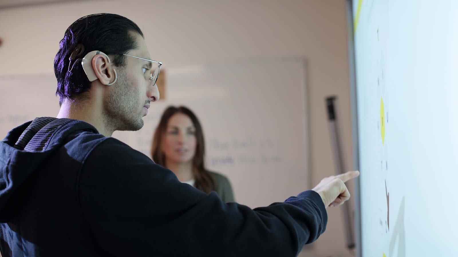 deaf and hard of hearing student wearing a hearing aid is pointing at a white board while an instructor is looking.