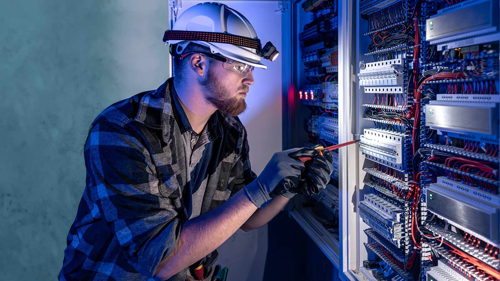 A male electrician, wearing a hard hat with a headlamp, safety glasses, and gloves, uses a screwdriver to work on an open electrical switchboard panel.