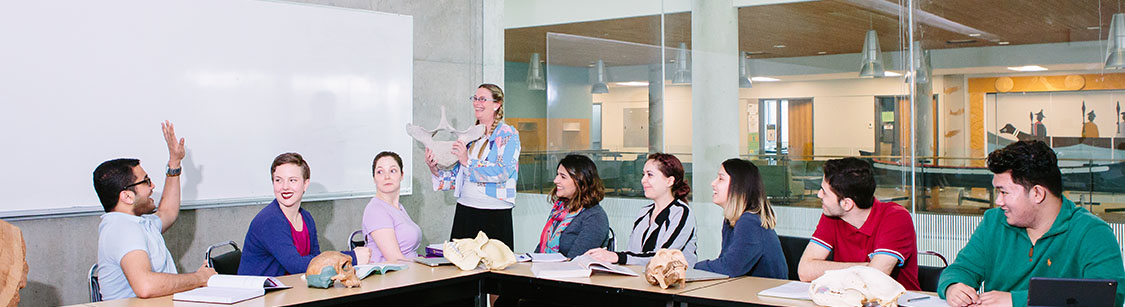 Students sitting in a classroom looking at a student who is rising his hand. A teacher is standing next to them smiling.